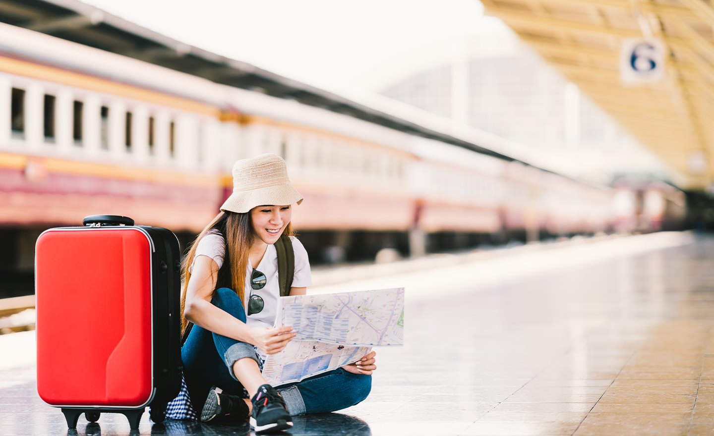 Young woman wearing hat sits on train platform with train behind her. She's looking at a map with her travel bag on the platform beside her.