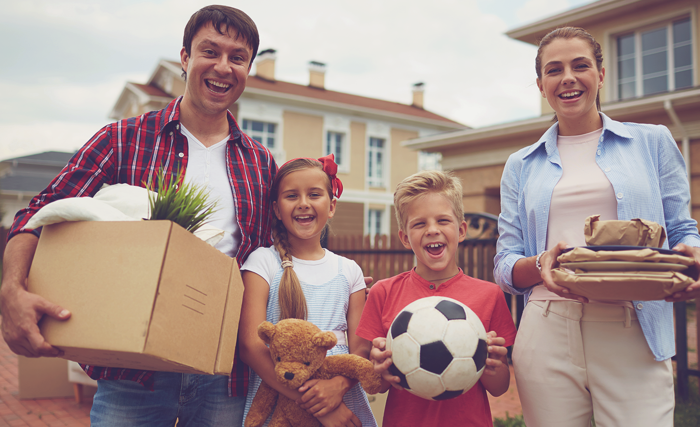 Young husband and wife couple with two children, a boy and a girl, grin widely while holding boxes in front of their new house