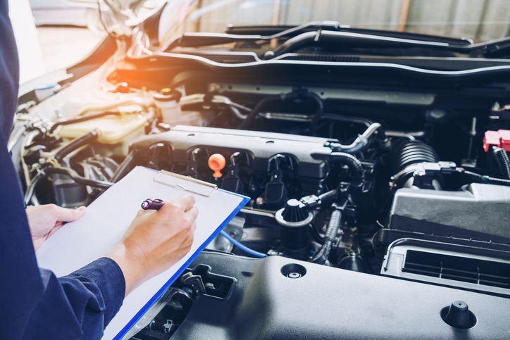 Mechanic looks under the hood of a car, taking notes of what needs to be done.