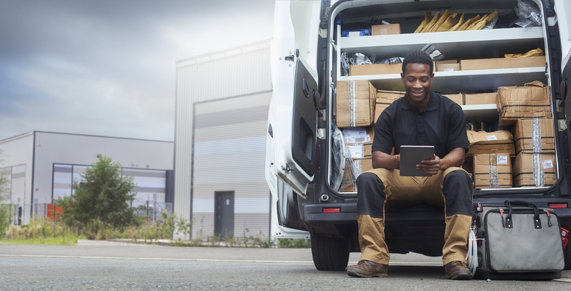 Young man sitting on the back bumper of his van looking at a tablet