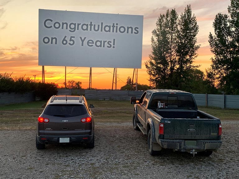 Two vehicles in front of a large drive-in movie theatre screen with the message Congratulations on 65 years
