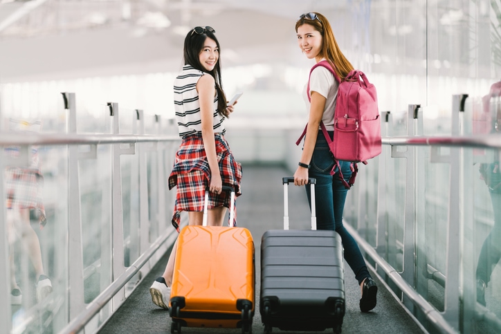 Two girls on moving walkway in airport carrying luggage on wheels look behind them at photographer