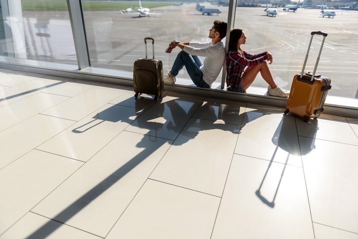 Couple sits back-to-back against glass in the airport waiting for their trip