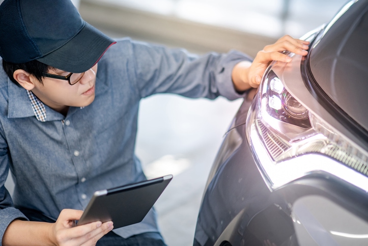 Young mechanic with digital tablet held in his hand inspects vehicle headlight in a service garage